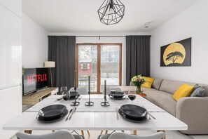 Una sala de estar moderna con comedor, un sofá con cojines amarillos, un cuadro de un árbol y una luna, y un televisor que muestra el logotipo de Netflix.