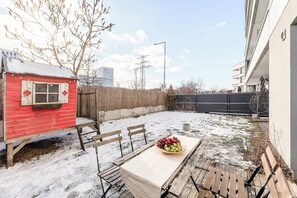 A snow-covered outdoor seating area with a table and chairs, a red wooden structure, and a building in the background. - Family Flat With Garden (Warsaw)