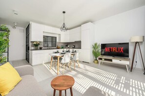 Eine moderne Küche mit weißen Schränken, eine Essecke mit Tisch und Stühlen, ein Sofa, ein Couchtisch und ein Fernseher, der 'NETFLIX' anzeigt.