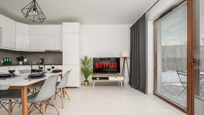Uma sala de estar moderna com área de jantar, uma TV exibindo 'NETFLIX' e uma janela grande com persianas.