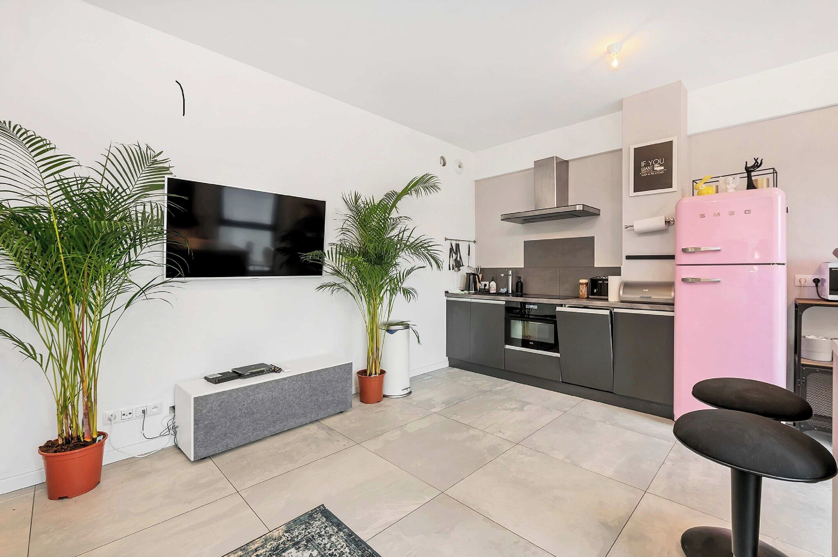 A modern kitchen with a flat-screen TV, a pink fridge, and two potted plants.