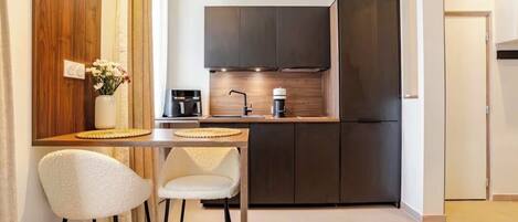 A compact kitchen with dark cabinets, a refrigerator, and a coffee maker. There is a small dining area with two chairs and a table.