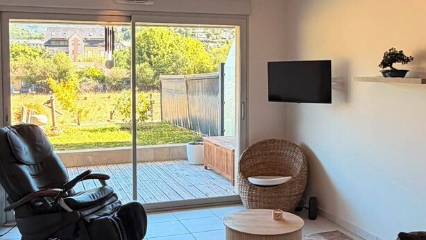 Ein Wohnzimmer mit einer Schiebetür zum Balkon, einem schwarzen Liegestuhl, einem runden Korbtisch und einem flachbildfernseher an der Wand.