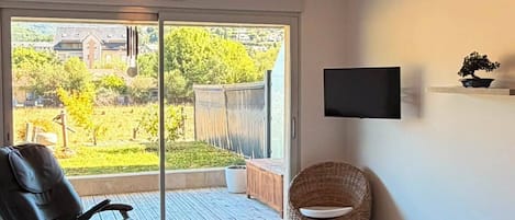 Una sala de estar con puerta corrediza de vidrio que da a un balcón, un sillón reclinable negro, una mesa redonda de mimbre y un televisor de pantalla plana montado en la pared.