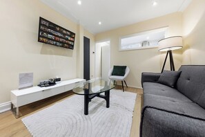 Ein modernes Wohnzimmer mit einem Glastisch, einem grauen Sofa, einem weißen Konsolentisch, einer schwarzen Stehlampe und einem an der Wand montierten Fernseher, der einen Filmplakat zeigt.