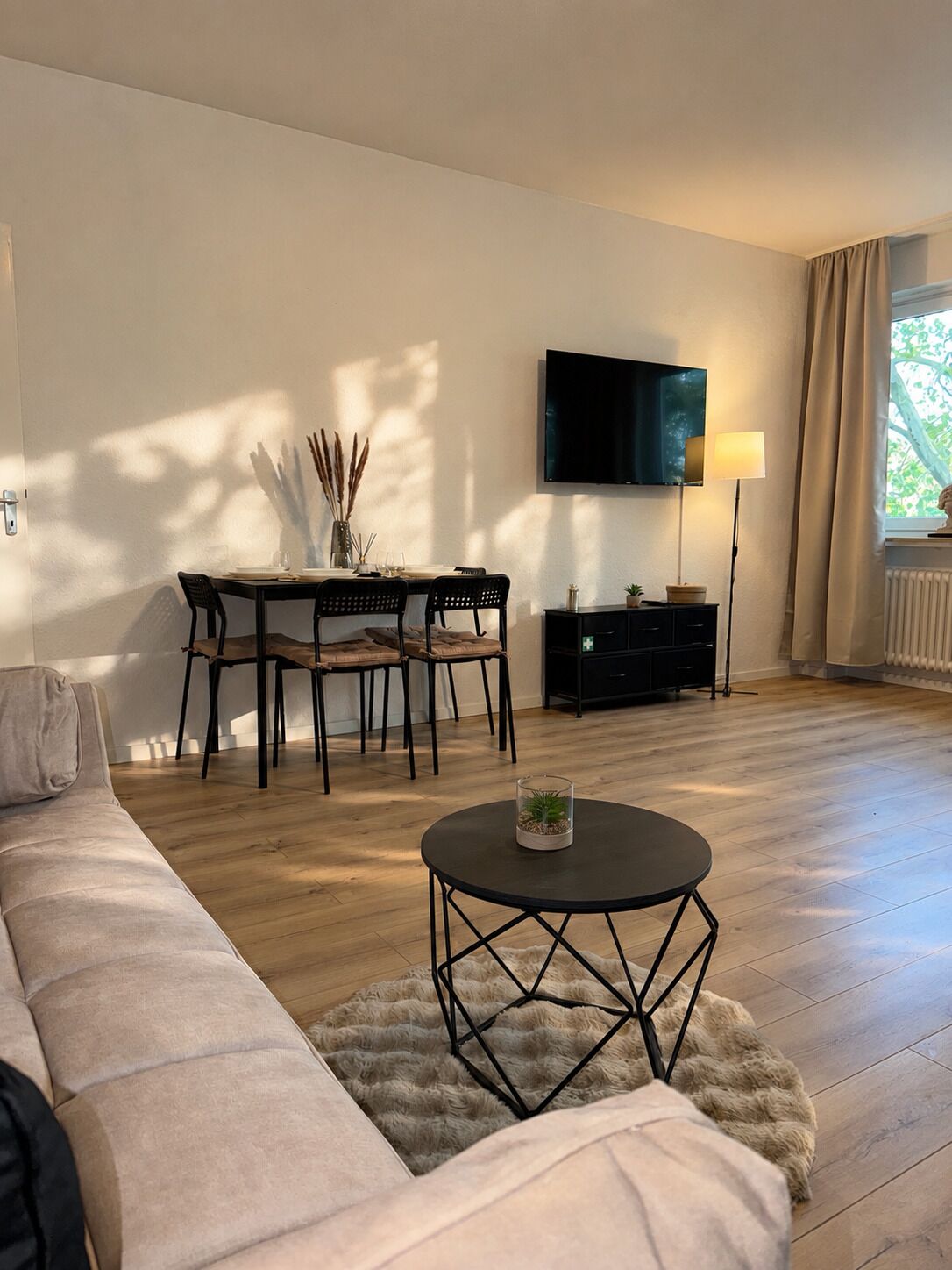 A living room with a sofa, a dining table with chairs, a TV mounted on the wall, a lamp, and a window with curtains.