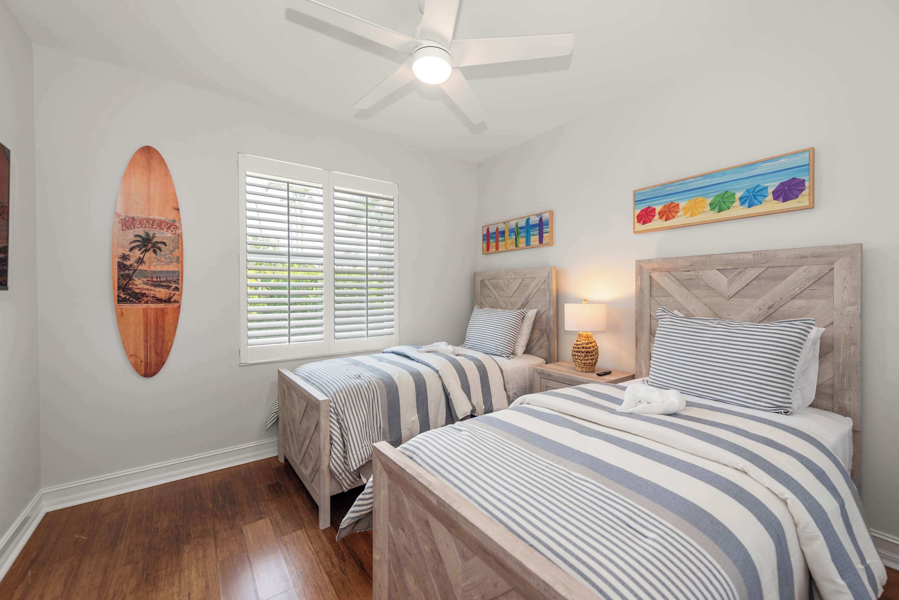 A bedroom with two beds, a ceiling fan, a beach-themed surfboard, and a beach umbrella painting.