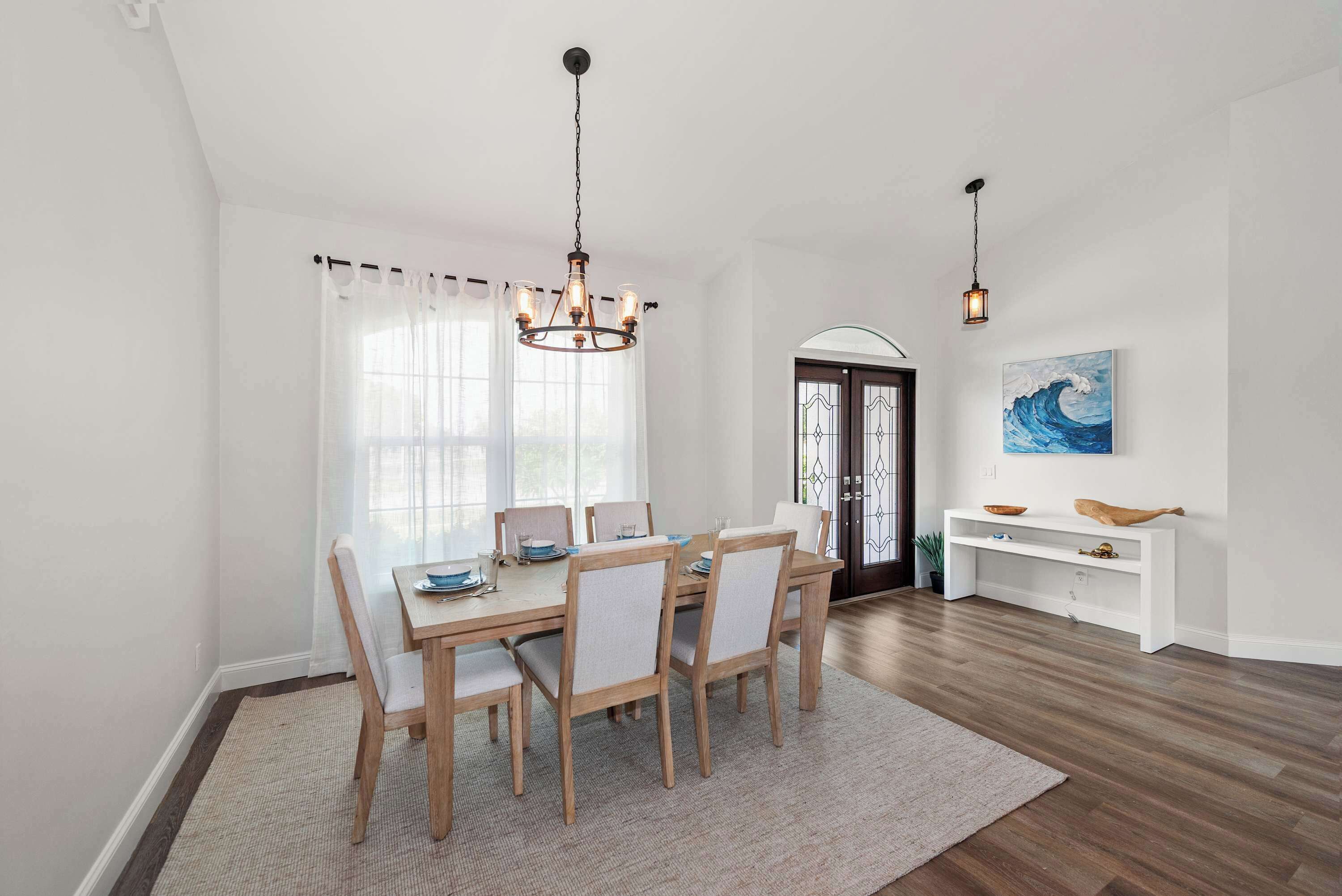 A dining room with a wooden table and chairs, a chandelier, and a view of the outdoors through large windows.
