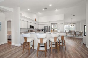 Een moderne keuken met witte kastjes, een centraal eiland met barstoelen en een eetgedeelte met een kroonluchter.
