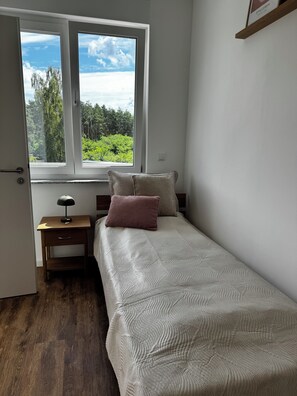 Un dormitorio con cama, mesita de noche, ventana y vistas al verdor, además de una puerta.