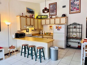 A kitchen with a central island, bar stools, a refrigerator with a sticker, and a shelf with glassware. - The Doak Lookout - 2BR Condo Overlooking Stadium and Minutes from Campus (Tallahassee)