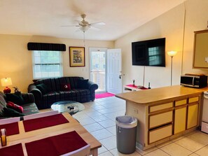 A compact living space with a sofa, a small coffee table, a TV, and a kitchenette area. - The Doak Lookout - 2BR Condo Overlooking Stadium and Minutes from Campus (Tallahassee)