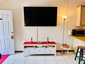 A room with a flat-screen TV mounted on the wall, a white cabinet with drawers, a small round table with a lamp, and a microwave on a shelf. - The Doak Lookout - 2BR Condo Overlooking Stadium and Minutes from Campus (Tallahassee)