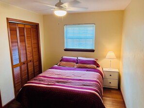 A bedroom with a bed, a nightstand, a lamp, a ceiling fan, a window with blinds, and a wooden closet. - The Doak Lookout - 2BR Condo Overlooking Stadium and Minutes from Campus (Tallahassee)