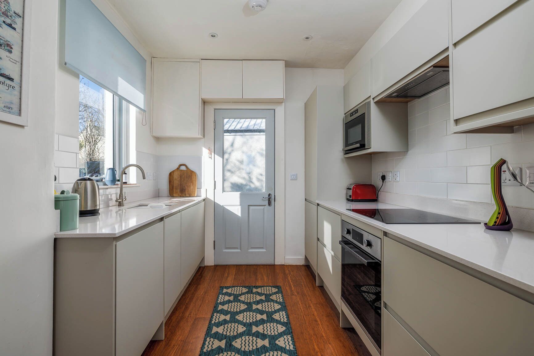 A modern kitchen with white cabinetry, a stainless steel kettle, a red toaster, and a patterned rug.