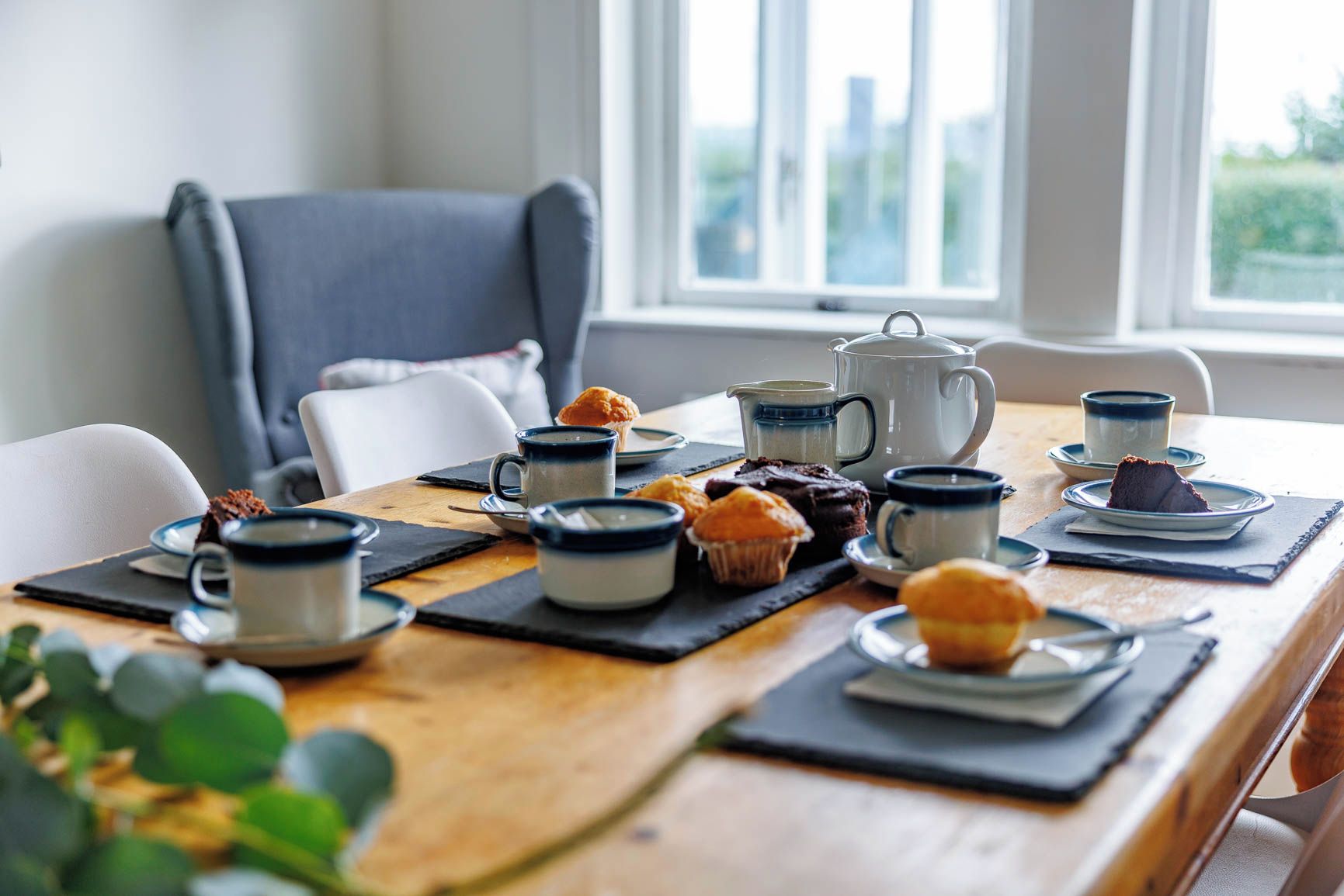 A dining table with a coffee pot, cups, and muffins, set in a room with a window and a chair.