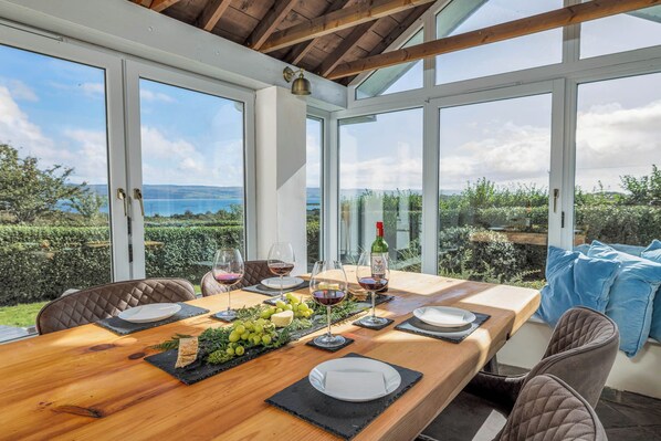 A dining area with a wooden table set for two, surrounded by chairs, with a view of a vineyard and mountains through large windows.
