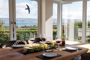 A dining table set with wine glasses, a bottle of wine, and a fruit platter, overlooking a scenic view with a bird silhouette.