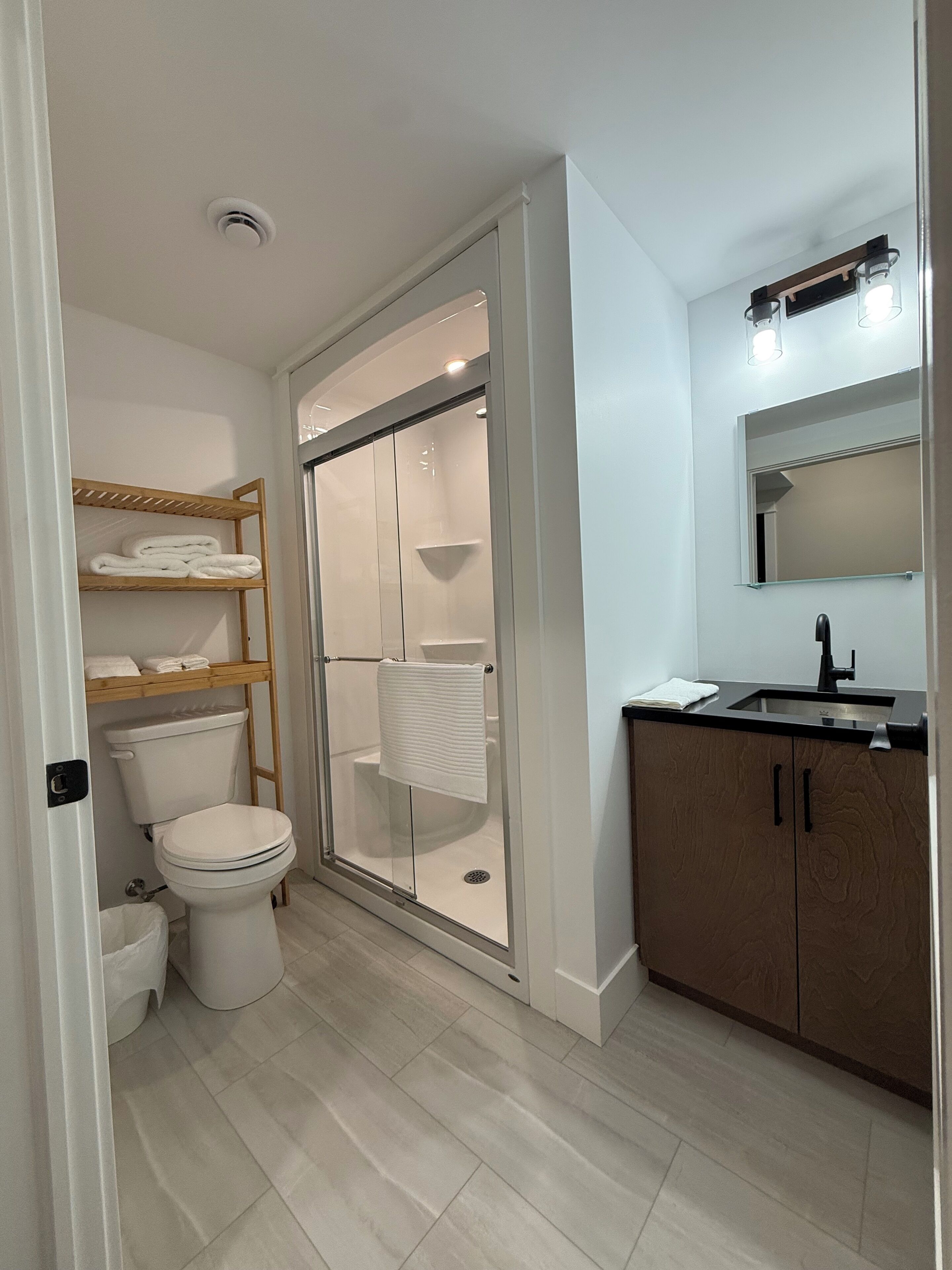 A modern bathroom with a toilet, a glass-enclosed shower, a wooden shelf with towels, and a sink with a mirror.