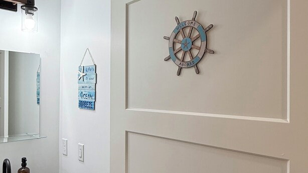 A bathroom with a wooden cabinet, a mirror, a ship's wheel decoration, and a welcome sign. - Sakura Valley Studio Suite #10 / Private Entrance in beautiful Annapolis Valley (North Alton)