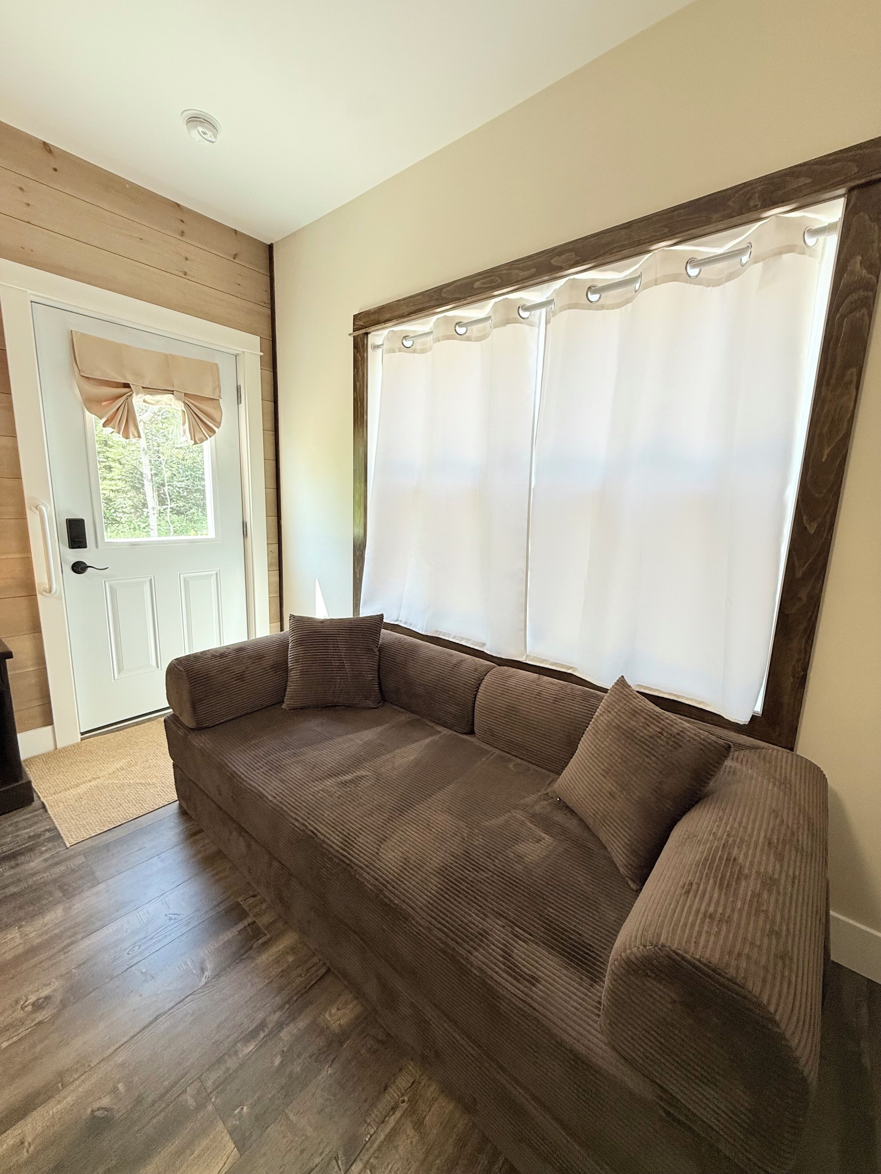 Ein Zimmer mit Holzboden, einem braunen Sofa, einer weißen Tür mit beigem Vorhang und einem Fenster mit weißen Vorhängen.