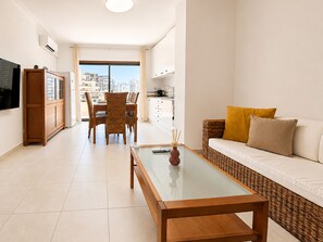 A modern living room with a glass-top coffee table, a sofa with cushions, a dining area with wicker chairs, and a kitchen in the background. - Apartment Rocha | Sea View & Pool | Pulse (Praia da Rocha)