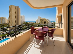 A balcony with a table and chairs, overlooking a swimming pool and high-rise buildings. - Apartment Rocha | Sea View & Pool | Pulse (Praia da Rocha)