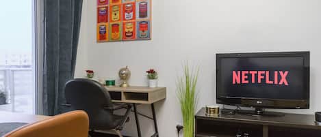 Una habitación con una televisión mostrando 'NETFLIX', un escritorio con un globo terráqueo, una silla, una planta en maceta y un cuadro con temática de latas.