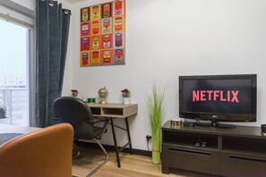 Ein Zimmer mit Fernseher, der 'NETFLIX' anzeigt, einem Schreibtisch mit Globus, einem Sessel, einer Topfpflanze und einem Wandbild mit Motiv aus Konservendosen.