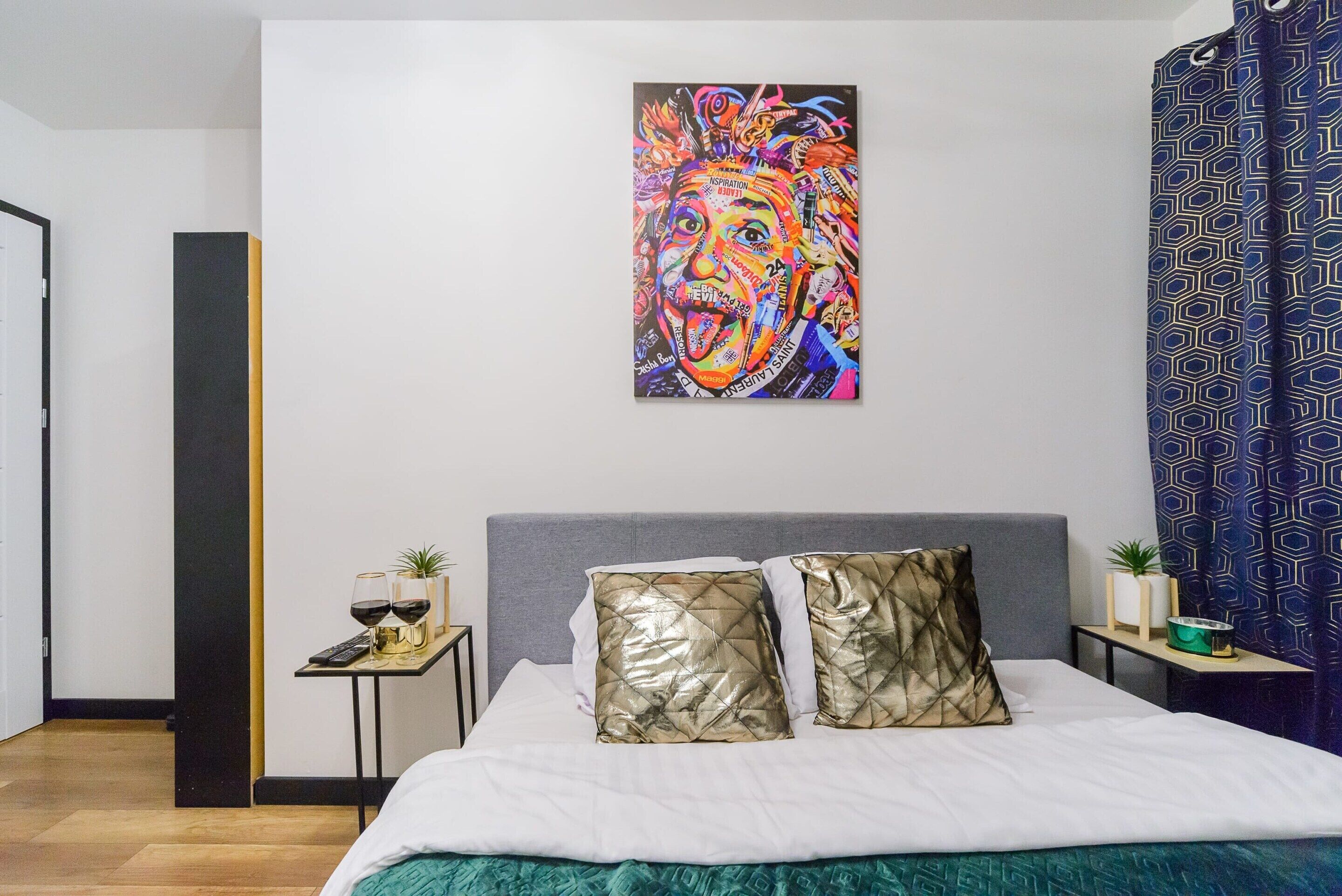 Un dormitorio con cama, dos almohadas, una mesita de noche, un cuadro y una cortina estampada.