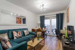 Ein modernes Wohnzimmer mit einem türkisfarbenen Sofa, einem Couchtisch mit Weingläsern, einem Fernseher und einem Essbereich mit Stühlen und Tisch.