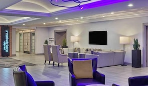 A modern hotel lobby with seating areas, a reception desk, and a television screen.