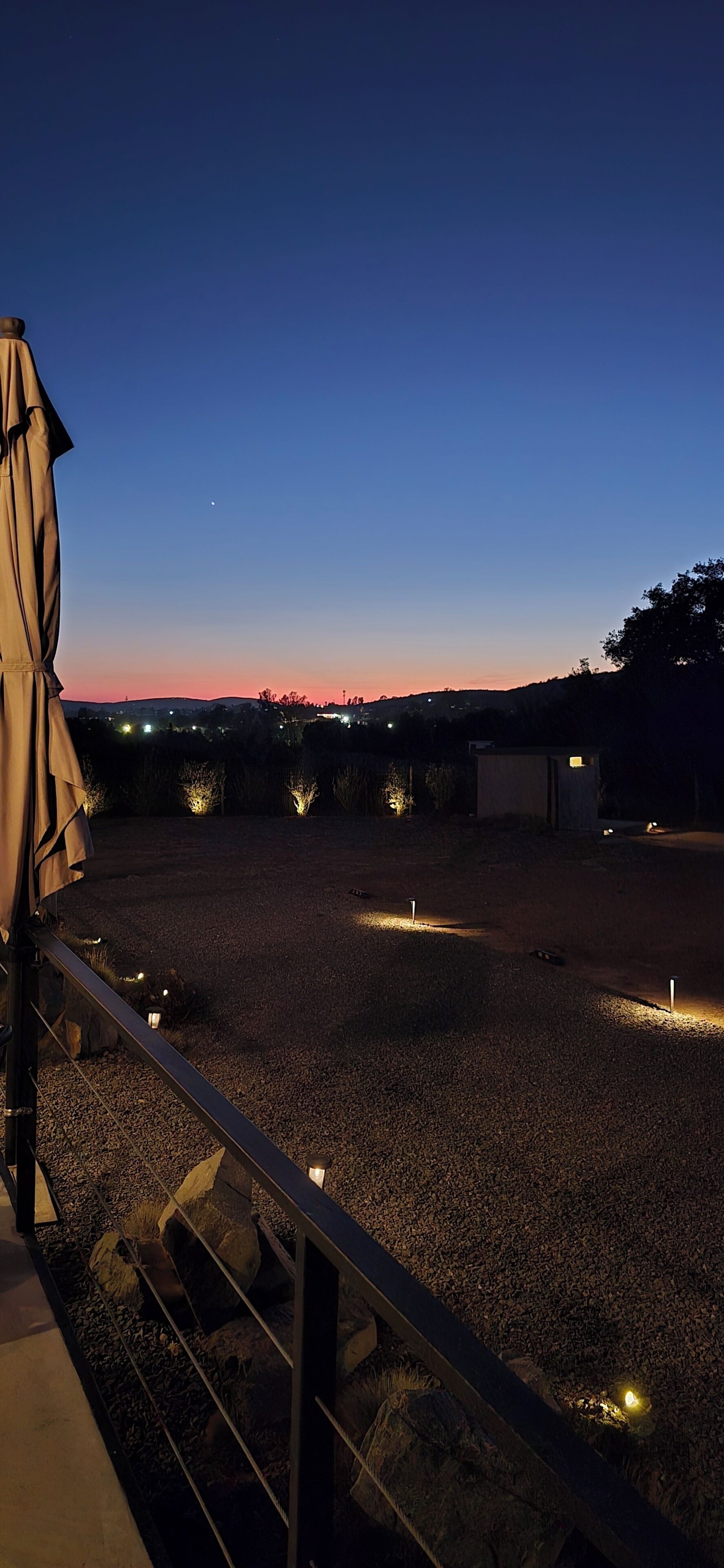 Uma área externa com piso de cascalho, guarda-corpo de metal e algumas luzes iluminadas.