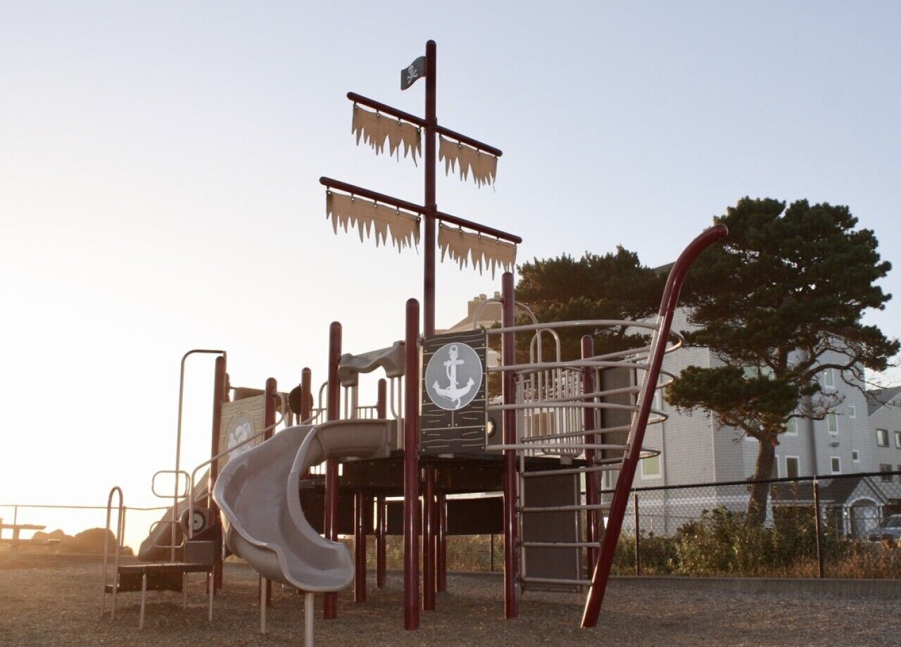 Un parque infantil con tobogán y estructuras de escalada, con árboles y edificios como fondo.