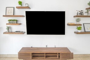 A flat-screen TV mounted on a white wall with wooden shelves above and below it, holding decorative items and plants. - Casa de Arizona: 3 BR Luxury Stay + King Bed (Gilbert)