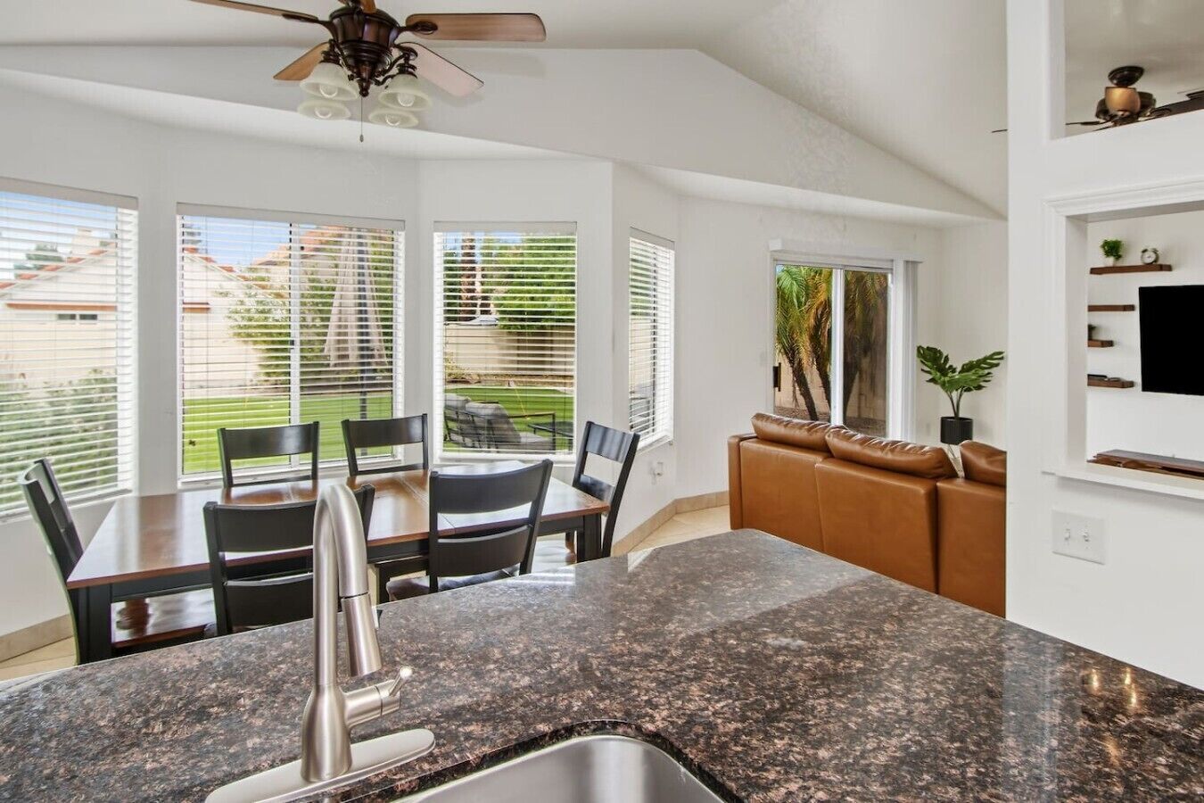 A kitchen with granite countertops, a dining table with chairs, a ceiling fan, and a view of the outdoors through large windows.