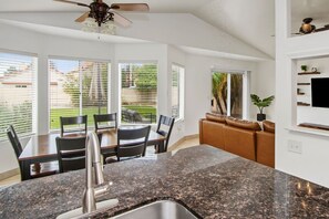 A kitchen with granite countertops, a dining table with chairs, a ceiling fan, and a view of the outdoors through large windows. - Casa de Arizona: 3 BR Luxury Stay + King Bed (Gilbert)