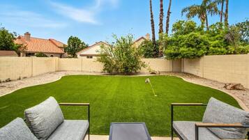 A backyard with a lawn, a putting green, and a patio area with seating.