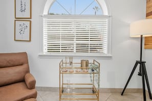 A room with a brown chair, a window with white blinds, a floor lamp, and a shelf with decorative items. - Casa de Arizona: 3 BR Luxury Stay + King Bed (Gilbert)