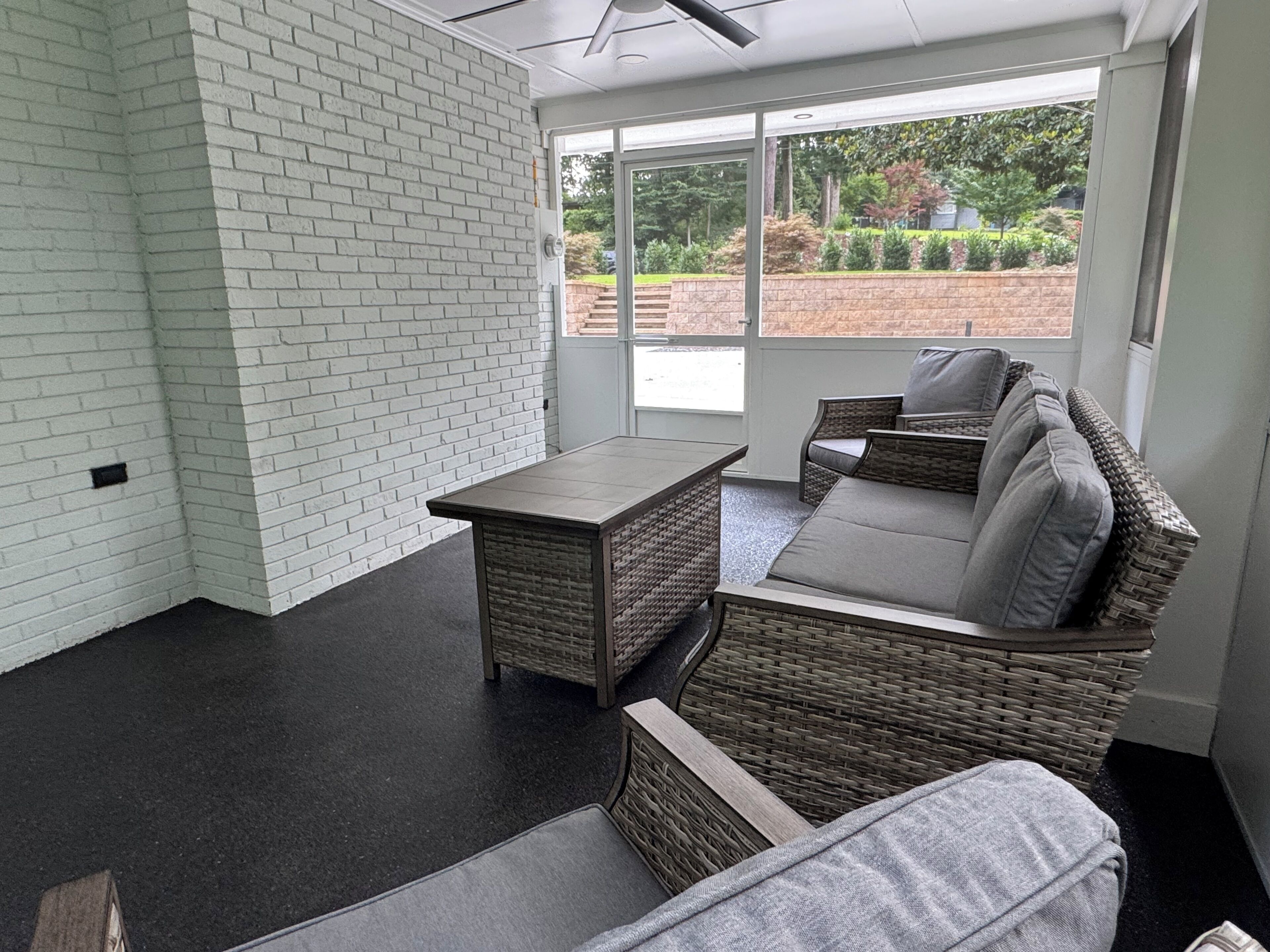 A modern interior with a white brick wall, a dark floor, wicker furniture, and a large window offering a view of greenery.