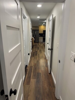 Un couloir avec des portes blanches, un coin cuisine équipé d’un réfrigérateur et d’un four à micro-ondes, et un plancher en bois.