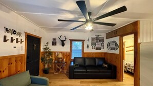 A cozy living room with a black leather sofa, a blue armchair, a wooden ceiling fan, and a black furry rug. - Alpine Retreat -Hot Tub • Walk to Village, Lake (Big Bear Lake)