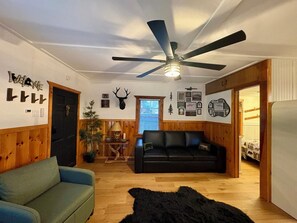 Een gezellige woonkamer met een zwarte leren bank, een blauwe fauteuil, een houten plafondventilator en een donzig zwart vloerkleed.