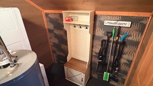 A compact storage area with a shelf holding cleaning supplies, a blue barrel, and a door with a 'mudroom' sign. - Alpine Retreat -Hot Tub • Walk to Village, Lake (Big Bear Lake)