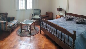 Una habitació amb un terra de fusta, un llit amb edredó blau, una cadira, una taula i una finestra amb persianes.