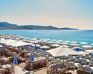 A beachside restaurant with outdoor seating and umbrellas, overlooking a calm sea and distant hills. - 3-minute walk to the beach, private villa, garden, parking, tram (Nice)