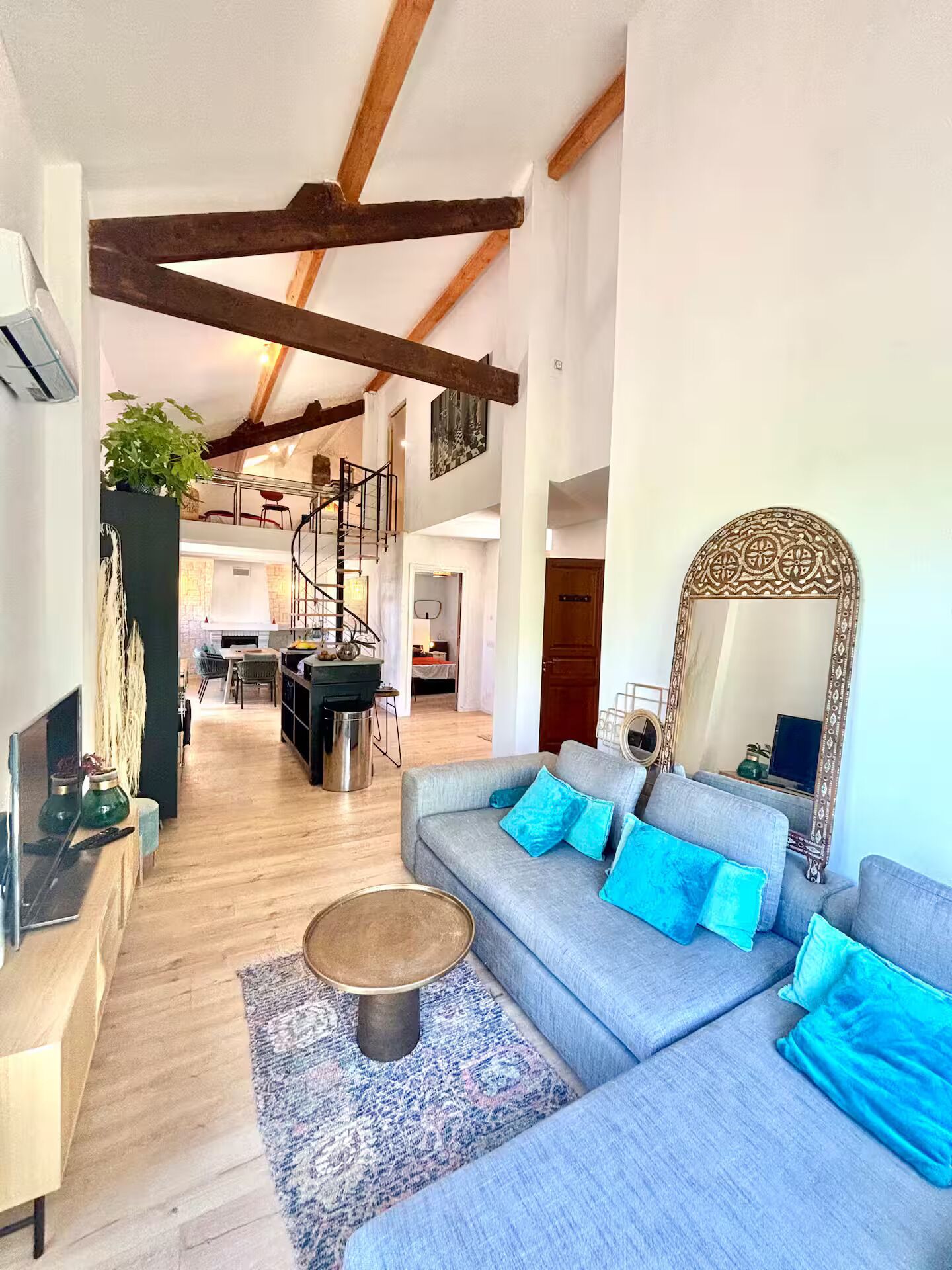 A spacious living area with a large sofa, a coffee table, and a decorative mirror. A spiral staircase is visible in the background, leading to an upper level.