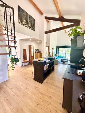 Ein geräumiges Wohnzimmer mit einer Holztreppe, eine Essecke mit einer zentralen Kochinsel und ein großes Fenster mit Blick ins Freie.