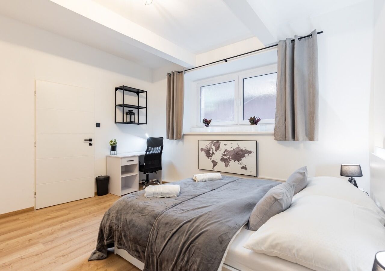 Een slaapkamer met een bed, een bureau, een stoel, een raam met gordijnen, een wereldkaart als kunstwerk en een kleine plant.