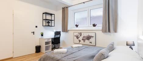 A bedroom with a bed, a desk, a chair, a window with curtains, a world map artwork, and a small plant.
